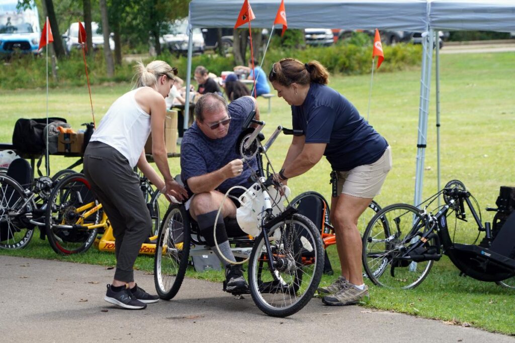 Man is being assisted by 2 staff to use a tricycle with hand brakes. Different accessible bikes are in the background.