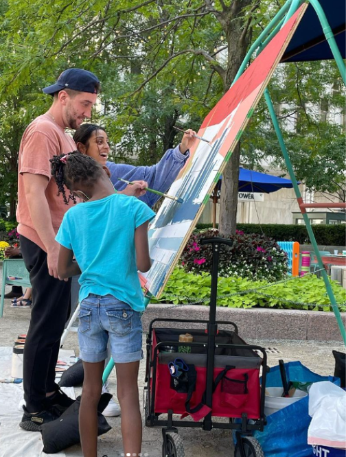 Three people are painting on a large canvas in a park.